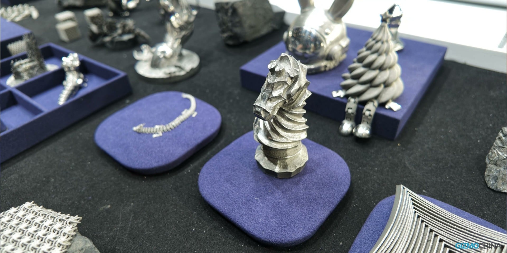 Various metal 3D printed objects displayed on blue pads at a trade show booth.